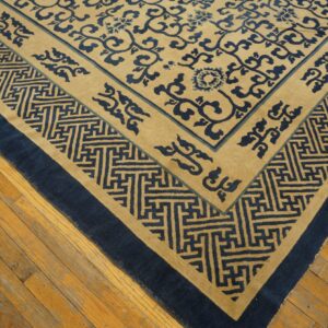 Tan and dark blue rug featuring floral scrollwork and geometric key borders on worn wood flooring.
