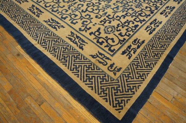 Tan and dark blue rug featuring floral scrollwork and geometric key borders on worn wood flooring.