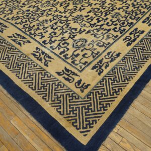 Tan rug with dark blue floral and meander patterns shown on distressed wooden floorboards.