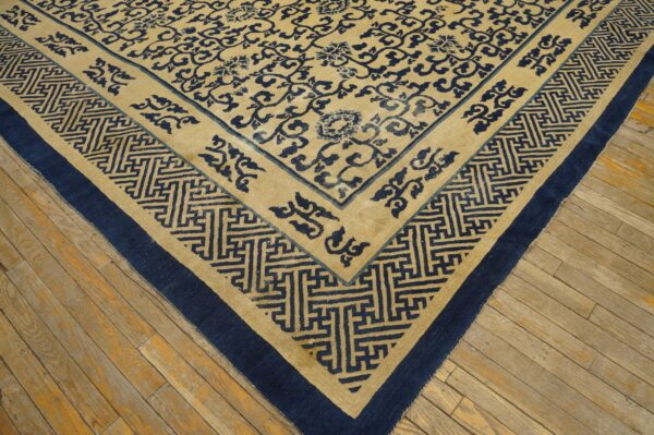 Tan rug with dark blue floral and meander patterns shown on distressed wooden floorboards.