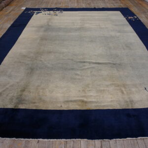 A light beige rug with a wide dark blue border and corner floral motifs rests on worn wood flooring.