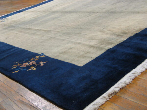 Low-pile beige and navy rug with a floral border accent lying on worn wood planks.