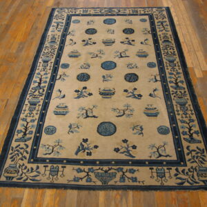 Tan rug with detailed blue borders and center motifs of vases, branches, and medallions rests on plank flooring.
