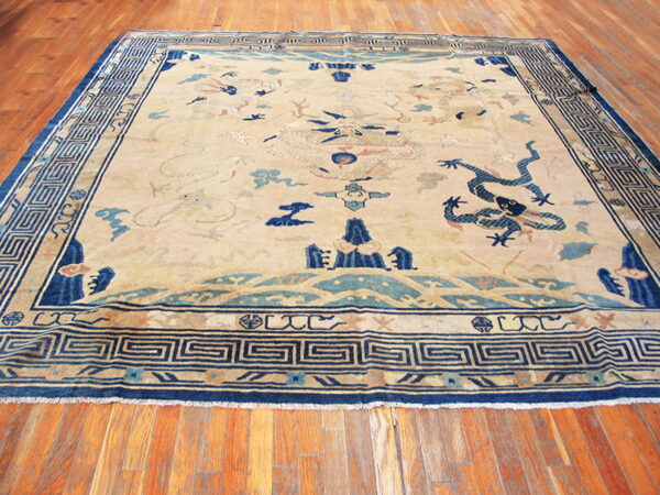 Tan rug with blue dragon patterns and geometric borders laying on shiny reddish-brown hardwood.