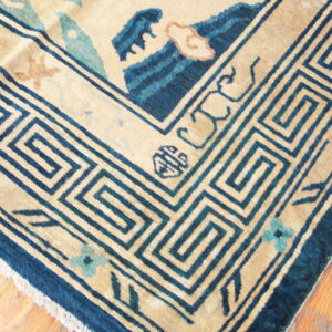 Low-pile beige rug with a dark blue key border and teal floral accents rests on rustic wood flooring.