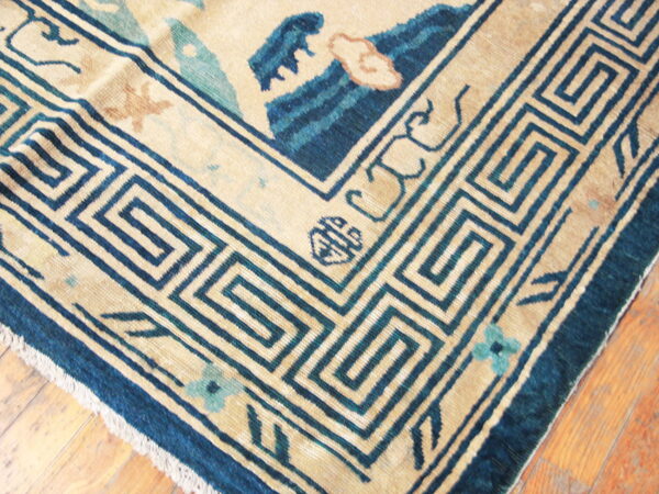 Low-pile beige rug with a dark blue key border and teal floral accents rests on rustic wood flooring.