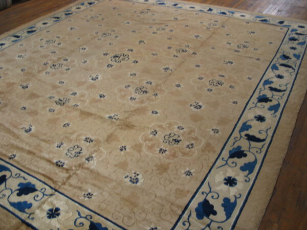 Beige low-pile rug featuring scattered blue floral motifs and a wide blue and tan stylized border over hardwood flooring.