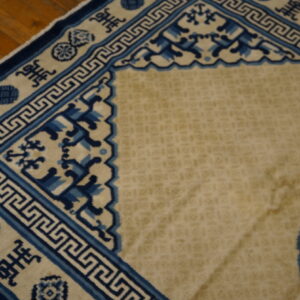 Tan rug with complex blue Greek key and symbol borders lying on warm brown wood flooring.