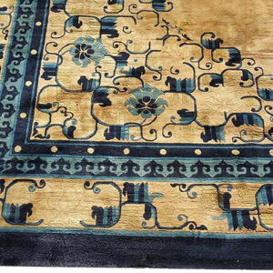 A rug corner showing a tan field and elaborate navy and teal geometric scrollwork borders.