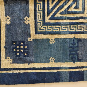 Close-up of a blue rug with geometric beige borders and stepped patterns lying on a pale floor.