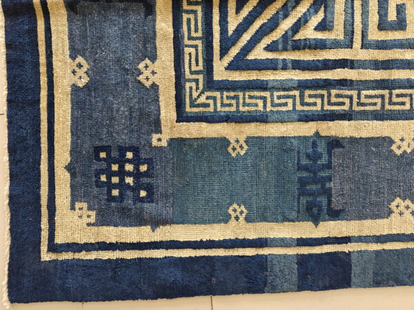 Antique chinese - peking chinese knotted rug 5 Close-up of a blue rug with geometric beige borders and stepped patterns lying on a pale floor.