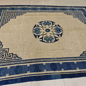 A tan and dark blue rug featuring a geometric border and round central medallion, displayed on white tile.