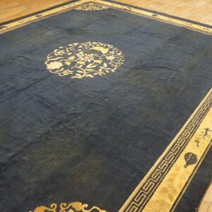 Dark blue area rug with a yellow floral medallion and fretwork border rests on hardwood planks.