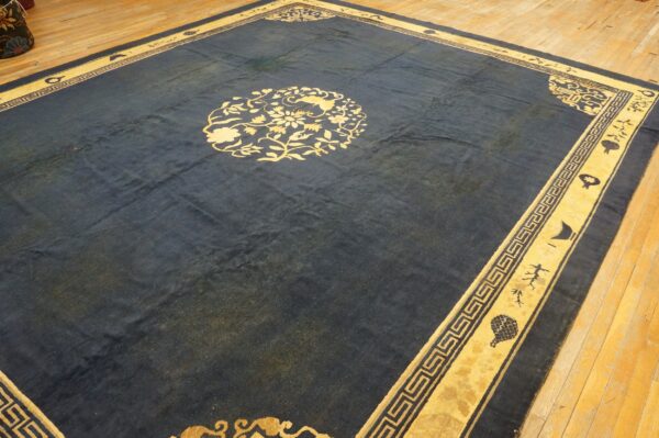 Dark blue area rug with a yellow floral medallion and fretwork border rests on hardwood planks.