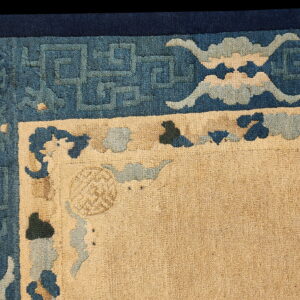 Close-up of a beige rug corner bordered by dark blue geometric fretwork and stylized cloud motifs.
