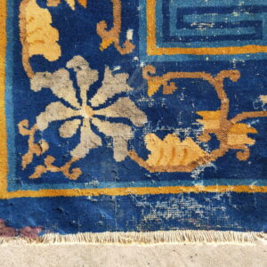 Close-up of a distressed royal blue rug featuring gold and grey floral motifs and white fringe on cement.