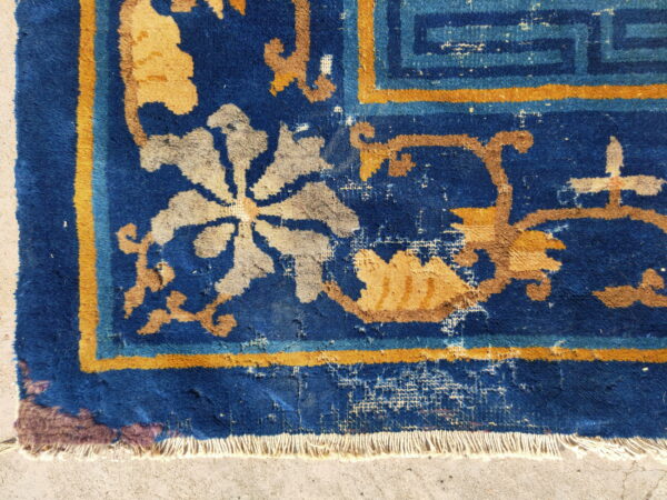 Close-up of a distressed royal blue rug featuring gold and grey floral motifs and white fringe on cement.