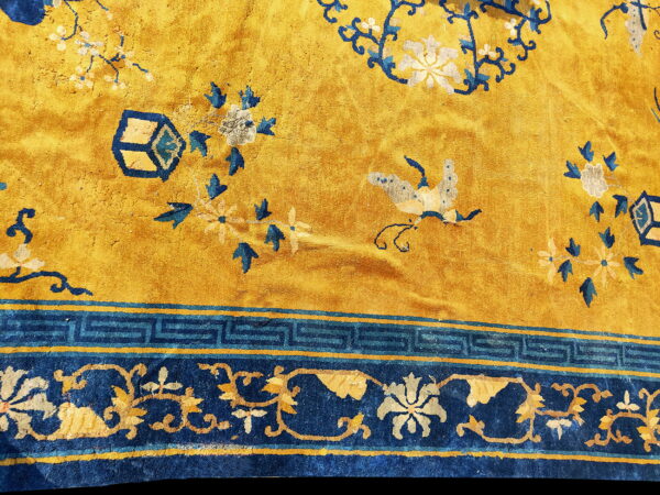 Gold and blue rug detail showing central floral motifs and a border with greek key and scrolling patterns.