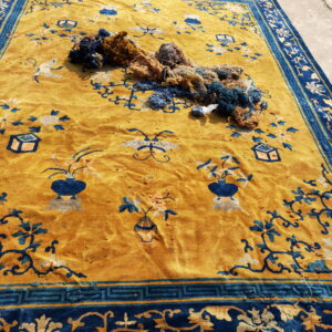 Worn golden yellow rug with blue stylized floral motifs sits on concrete with repair yarn.
