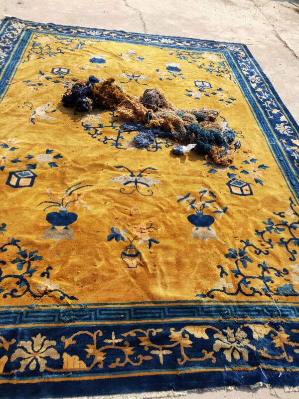 Worn golden yellow rug with blue stylized floral motifs sits on concrete with repair yarn.