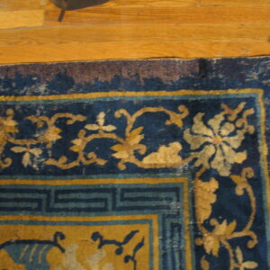 Worn blue and gold rug displaying a floral and fret border pattern sits on a light hardwood floor.