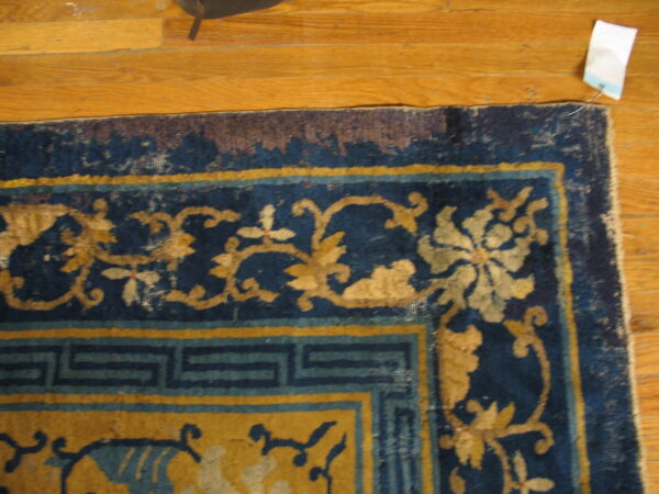 Worn blue and gold rug displaying a floral and fret border pattern sits on a light hardwood floor.