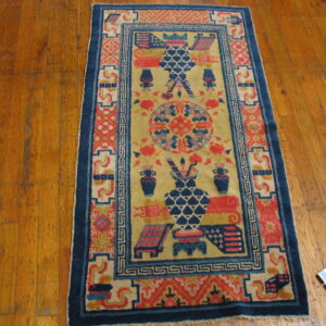 Small, patterned rug in yellow, coral, and navy blue with various objects rests on distressed wood flooring.
