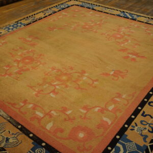 A gold low-pile rug with pink floral designs and geometric borders in blue and black on a wood floor.