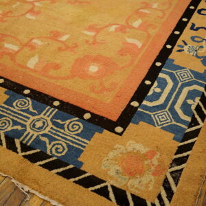 Corner detail of a tan rug featuring borders in black, blue, pink, and cream geometric and floral designs on wood flooring.