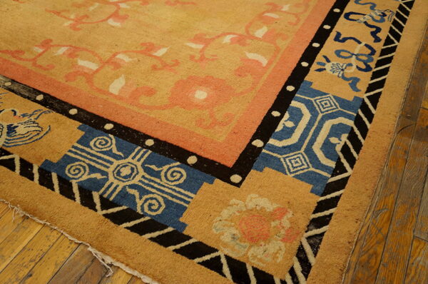 Corner detail of a tan rug featuring borders in black, blue, pink, and cream geometric and floral designs on wood flooring.