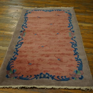 Muted coral rug with a blue scrolling border and taupe edge rests on distressed wood planks.