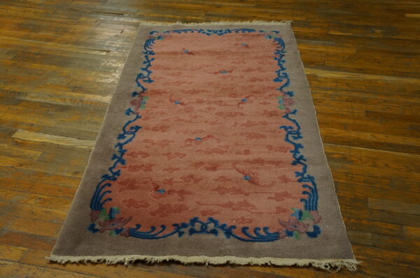 Muted coral rug with a blue scrolling border and taupe edge rests on distressed wood planks.