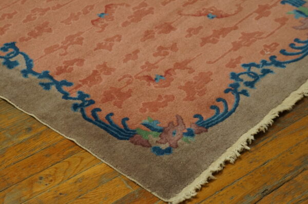Pink and salmon patterned rug corner with a scrolling blue and multicolored floral border on wood floor.