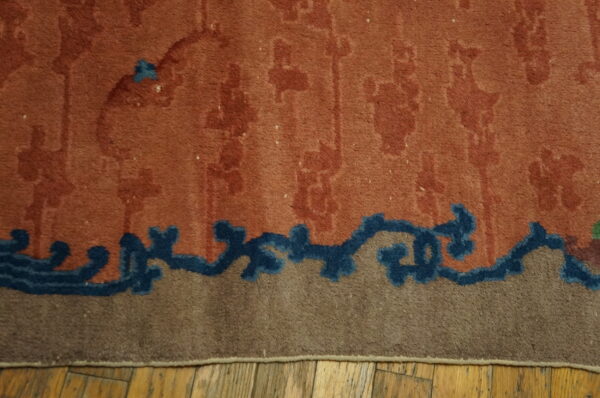 Burnt orange rug features subtle motifs above a dark blue border section and tan edge on exposed wood planks.