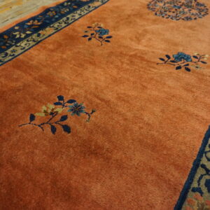 High-pile orange rug displaying blue floral motifs and a detailed blue and beige patterned border on wood flooring.