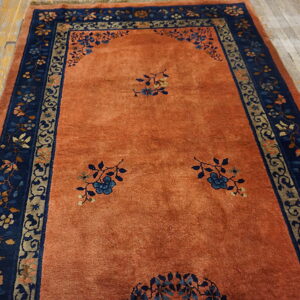 Terracotta-orange rug featuring blue floral sprigs and a detailed navy and beige border on light wood floors.