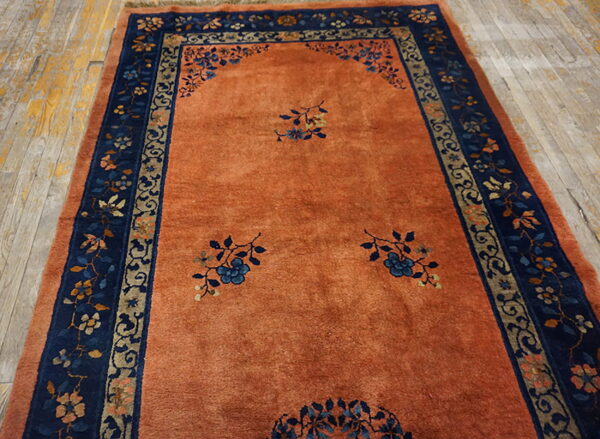 Terracotta-orange rug featuring blue floral sprigs and a detailed navy and beige border on light wood floors.