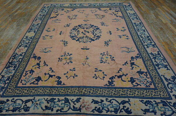 Low-pile pink rug with blue and yellow floral motifs and complex borders on a distressed wood floor.