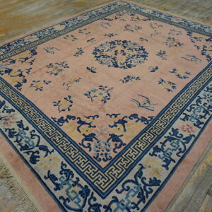 A large pink rug with blue botanical patterns and a complex geometric border on distressed wood flooring.