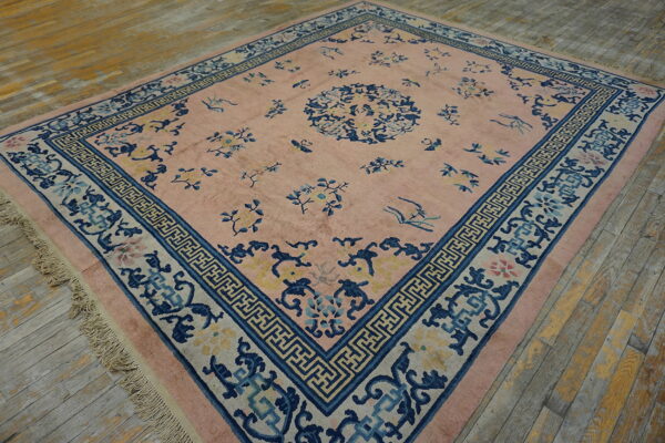 A large pink rug with blue botanical patterns and a complex geometric border on distressed wood flooring.