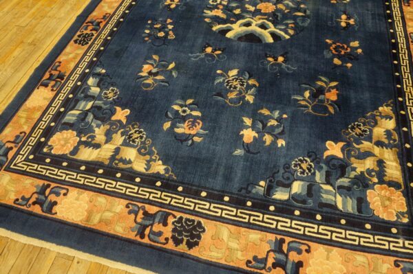Dark blue rug featuring gold and peach floral motifs and a greek key border over wood flooring.