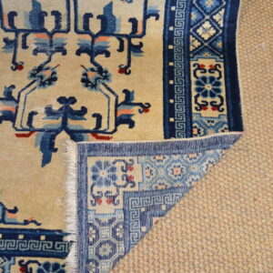 Beige rug with navy and blue floral motifs folded back, showing fringe over a textured tan rug.