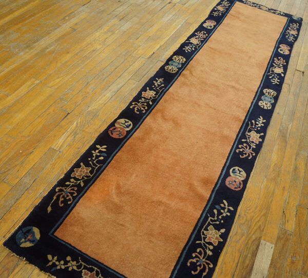A runner rug with a solid salmon field and a dark blue floral border rests on distressed wood flooring.