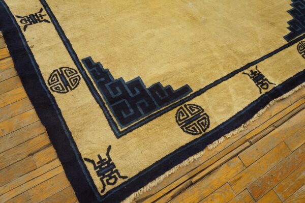 Gold rug corner with navy border showing geometric key patterns and symbols on rough wooden flooring.