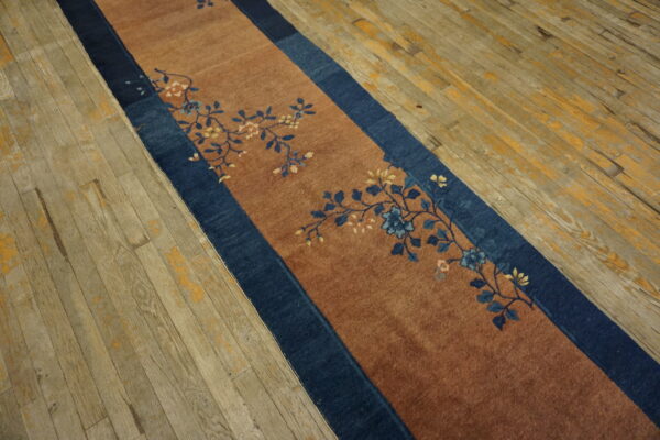 Rust runner rug with a navy border and blue floral pattern rests on gray and yellow distressed wood flooring.