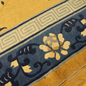 Gold rug with navy and cream floral patterns and a Greek key border resting on hardwood floors.
