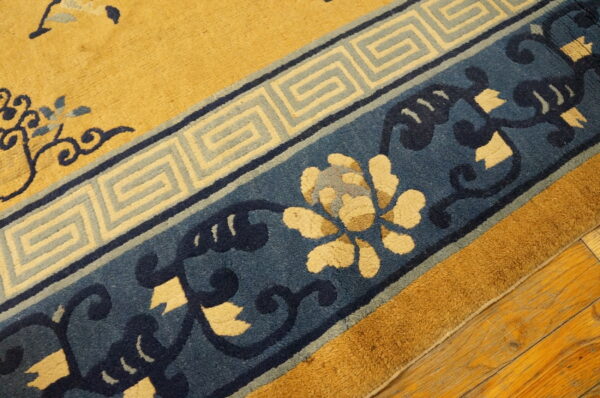 Gold rug with navy and cream floral patterns and a greek key border resting on hardwood floors.