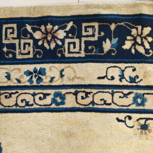 A high-pile rug detail showing navy blue borders with cream fretwork and teal floral motifs on a tan background.