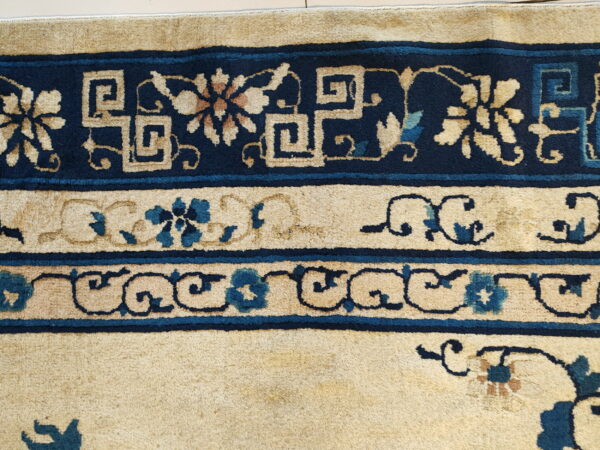 A high-pile rug detail showing navy blue borders with cream fretwork and teal floral motifs on a tan background.