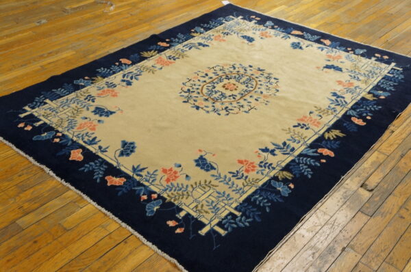 Beige and navy rug with blue, coral, and tan floral designs rests on a distressed wooden floor.